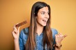 © Krakenimages.com - Young beautiful girl holding healthy protein bar standing over isolated yellow background pointing and showing with thumb up to the side with happy face smiling