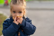 A Beautiful Little Caucasian Girl With Blond Hair And Eating Bread Eagerly With Her Hands Looks At The Camera With Sad Eyes