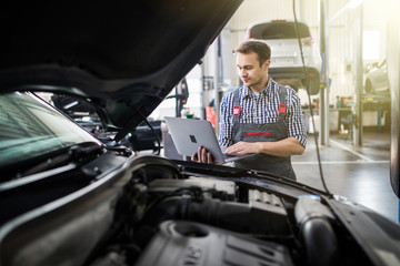 Wall Mural - Smiling young man mechanic using a laptop computer to check a car engine