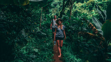 Couple Walking In A Green Jungle. Women With A Hat, Men With A Backpack In Tanzania Africa
