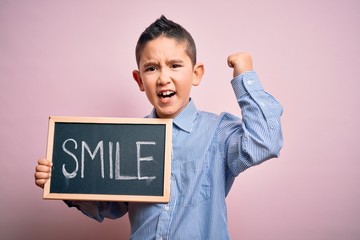 Poster - Young little boy kid showing blackboard with smile word as happy message over pink background annoyed and frustrated shouting with anger, crazy and yelling with raised hand, anger concept