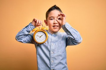 Young little boy kid holding classic bell alarm clock over isolated yellow background with happy face smiling doing ok sign with hand on eye looking through fingers