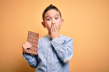 Young little boy kid eating sweet chocolate bar for dessert over isolated yellow background cover mouth with hand shocked with shame for mistake, expression of fear, scared in silence, secret concept