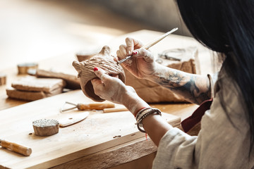 Craftsperson Concept. Young woman sitting at table making pottery indoors using modeling tool making shape back view close-up