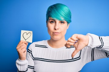 Sticker - Young woman with blue fashion hair holding romantic heart shape on note over isolated background with angry face, negative sign showing dislike with thumbs down, rejection concept