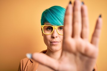 Canvas Print - Young beautiful woman with blue fashion hair wearing casual glasses over yellow background with open hand doing stop sign with serious and confident expression, defense gesture