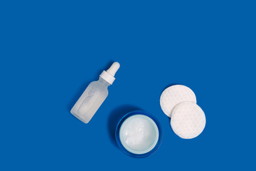 Glass eye dropper bottle with facial serum, jar of white moisturizing lotion, and round cotton pads on blue background / clean skin care routine still life