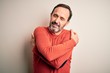 © Krakenimages.com - Middle age hoary man wearing casual orange sweater standing over isolated white background Hugging oneself happy and positive, smiling confident. Self love and self care