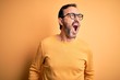 © Krakenimages.com - Middle age hoary man wearing casual sweater and glasses over isolated yellow background angry and mad screaming frustrated and furious, shouting with anger. Rage and aggressive concept.
