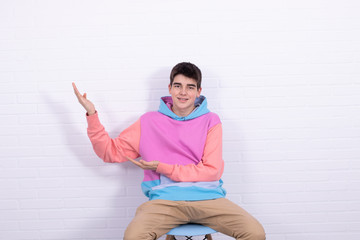 young man or teenager sitting on chair pointing at white brick wall background