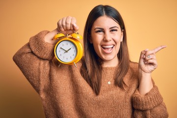 Poster - Young beautiful woman holding alarm clock standing over isolated orange background very happy pointing with hand and finger to the side