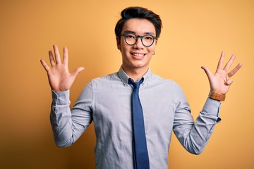 Young handsome chinese businessman wearing glasses and tie over yellow background showing and pointing up with fingers number nine while smiling confident and happy.