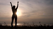 Back View Of Female Silhouette In Sportswear Raising Arms Up And Expressing Triumph While Standing On Coast At Sunset. Sporty Woman With Hands Up Meeting Setting Sun On Seashore Rejoicing Success