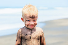 Funny Portrait Of Happy Smiling Baby With Dirty Face And Hands Have Fun On Sea Beach. Little Boy Playing With Black Sand. Active Kids Outdoor Leisure. Family Lifestyle On Summer Vacation With Children