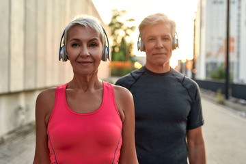 Wall Mural - Training with music. Portrait of happy and beautiful middle-aged couple in headphones looking at camera with smile while training together outdoors