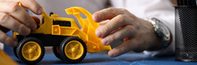 Close-up Of Businessman Hands Driving Toy Yellow Excavator. Builder Holding Backhoe Loader And Showing Future Restructuring Of Big City. Business And Construction Concept