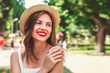 © Evgenia - A young girl in straw hat with a red lipstick and white dress walks in the park, smiles and looks away in the street, copy space. Cute girl looks away and smiles.