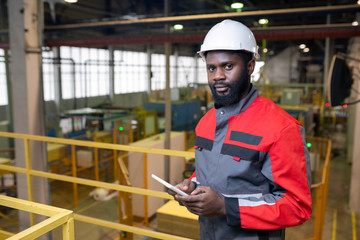 Portrait of confident bearded Black man in protective helmet using digital tablet at factory