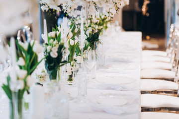 formal luxury elegant wedding decor restaurant tables served white tablecloth, plates, menus, glasses, tulips in vases, orchids, candles silver chairs, blue background