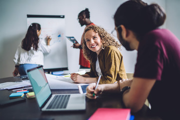 Wall Mural - Cheerful employees having nice conversation sitting at table while other diverse office workers using board to discuss project in workplace