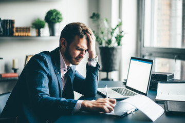 Manager sitting at the desk with laptop, reading important documents with puzzled expression and holding head with his hand.