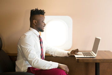 handsome african young man in white shirt in private jet cabin with laptop