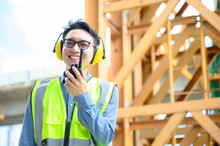 Young Asian Engineers Are Working On The Construction Site. Wear A Yellow Earmuff Sound Protection And Uniform Staff. Hand Holding Portable Radio Transceiver For Communication