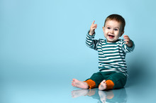 Little Baby Boy In Stylish Casual Clothing Barefoot Sitting On Floor And Smiling With Raised Hands Over Blue Wall Background