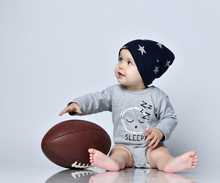 Little Baby Boy Toddler In Grey Casual Jumpsuit, Black Cap With Stars And Barefoot Sitting On Floor With Rugby Ball