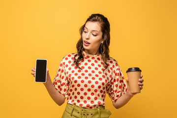 Wall Mural - attractive young woman holding coffee to go and smartphone with blank screen isolated on yellow