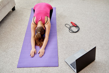 Wall Mural - Beautiful young woman doing stretching exercise on floor at home, online training on laptop computer, copy space. Full length portrait. Yoga, pilates, working out exercising