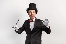 Excited Magician In Suit And Hat Holding Wand, Isolated On Grey