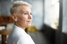 Close Up Profile Shot Of Elegant Short Haired Businesswoman Wearing Pearl Earrings And Stylish White Blouse Posing Isolated In Office Interior, Having Pensive Thoughtful Look. Job, Work And Mature Age