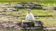 Little Caucasian Girl In A White Dress Climbs On The Old Stones Of The Ruins. Beautiful Elegant Child Walking In The Summer On The Meadow, Baby Girl Outdoor