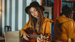 © Natalia Nieves/ADDICTIVE STOCK - Young stylish woman with dreadlocks wearing yellow coat and black hat sitting on old wooden table back to mirror and playing Hawaiian guitar ukulele in room with antique furniture