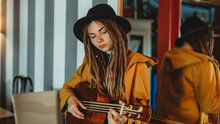Young Stylish Woman With Dreadlocks Wearing Yellow Coat And Black Hat Sitting On Old Wooden Table Back To Mirror And Playing Hawaiian Guitar Ukulele In Room With Antique Furniture