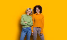 Portrait Of Two Happy Lovely Sisters One With Blonde Hair Another With Curly Hair Looking At Camera Laughing Isolated On Yellow Studio Wall.
