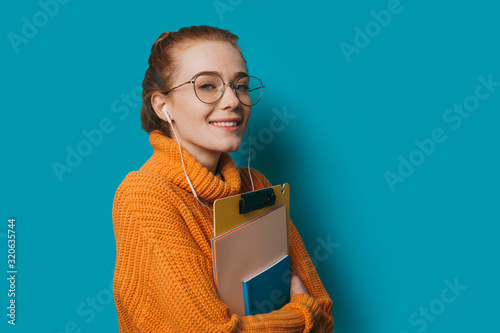 Fotomural Portrait of a young charming female student with red hair and freckles wearing glasses looking at camera smiling while holding her books against blue wall
