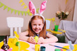 © Evgeniy Kalinovskiy - cheerful active child girl with red hair and rabbit ears paints an egg with yellow paint on a background of easter decor and smiles.Easter Traditions