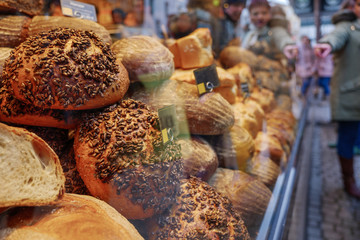 Store's display with heap and stack of wheat and rye breads with whole grain meal and rustic mustard crust at the front of bakery store on sidewalk with blur background of people. 