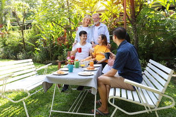 Happy multiethnic family sitting at a breakfast table in backyard outdoor on sunny day with smiling face. Happy caucasian and asian family sitting and having conversation with good time outdoor.