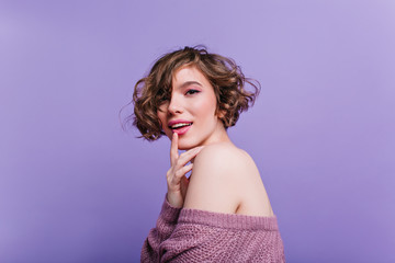 Wall Mural - Close-up portrait of lovable short-haired girl with pale skin posing on purple background. Indoor photo of gorgeous curly european lady playfully touching her lips.