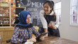 © PRPicturesProduction - Portrait of smiling young japanese waitress taking order from malay client in cafe bar writing note. muslim woman guest using digital tablet reading menu while friendly female barista introduce