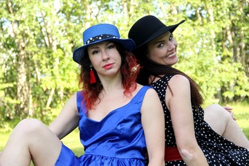 Two female girlfriends sit back-to-back on green lawn on a sunny day, close-up