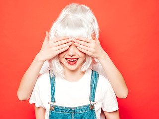 Two young sexy hipster girls in white wigs and red lips.Beautiful trendy women in summer clothes.Carefree models posing near red wall in studio.Covering her eyes and hugging from behind