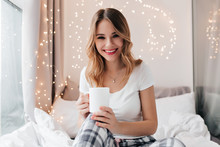 Wonderful European Girl With Trendy Necklace Sitting In Bed. Indoor Portrait Of Laughing Ecstatic Female Model Holding Cup Of Tea.