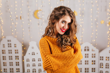 Wall Mural - Close-up portrait of gray-eyed girl with curly hair posing in knitted sweater on light background with toy houses