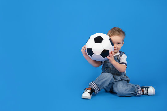 Сhild sits with a ball on a blue background and smiles, holding a soccer ball in front of him. Football competition. Sport concept of football, World Cup or European football tournament.