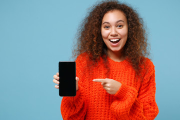 Excited african american girl in orange knitted clothes isolated on pastel blue background. People lifestyle concept. Mock up copy space. Pointing index finger on mobile phone with blank empty screen.