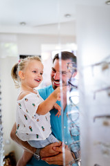 Wall Mural - Health care, eyesight and vision concept. Little girl choosing glasses with father at optics store